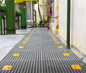 Rejilla plástica industrial instalada en planta de producción, con superficie antideslizante y señalización amarilla integrada para seguridad en pasarelas.